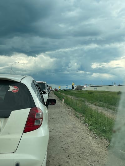This media file depicts a traffic jam on a cloudy day in Chita, Russia. The camera is positioned inside a white car, looking forward and slightly to the left. The scene is dominated by a dramatic sky filled with dark, heavy clouds, suggesting an impending storm or overcast weather. A line of cars stretches ahead, indicating a standstill or slow-moving traffic.
To the right of the road, there is a strip of green grass and a dirt path. In the distance, several signs and structures are visible, including a yellow and blue sign that appears to be for a gas station, and a triangular road sign indicating a hazard. There are also lampposts and what looks like a barrier or gate system further down the road. The overall impression is one of a mundane, everyday situation (a traffic jam) set against a visually striking sky.
There is no visible text that is clear enough to be read in detail. The time of day appears to be daytime, given the light, but the heavy cloud cover makes it difficult to determine the exact hour. The mood is neutral, characterized by the routine nature of a traffic jam and the somber atmosphere created by the weather. The setting suggests an outdoor, possibly semi-urban or highway location.