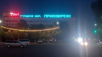 The image depicts a nighttime street scene in Priozersk, Kazakhstan. A large, multi-story building is illuminated by bright blue neon lights forming the text "ГУЛДЕНЕ БЕР, ПРИОЗЕРСК!". To the left of this text, a digital display shows "20:27" in red neon. A string of warm yellow lights hangs across the foreground, illuminating the trees below. Several vehicles are visible, including a silver station wagon in the left foreground, a white van behind it, and other vehicles further down the street. The street itself is dark, with the headlights of oncoming cars creating bright flares. Traffic signals are visible, with one showing a green light. The overall atmosphere is urban and nocturnal.