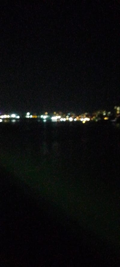 The image displays a nighttime scene, predominantly dark. A horizontal line of blurred, multi-colored lights, appearing as bokeh, stretches across the mid-ground. These lights are primarily white, yellow, and orange, with some green hues, suggesting a distant urban or residential area. The upper third of the image is a dark, indistinct sky, while the lower two-thirds are a dark, featureless expanse, likely water or unlit land. No individuals, specific structures, or discernible objects are visible. The scene is presented as a panoramic view of illuminated distant buildings or infrastructure in Junda ward, Kenya.