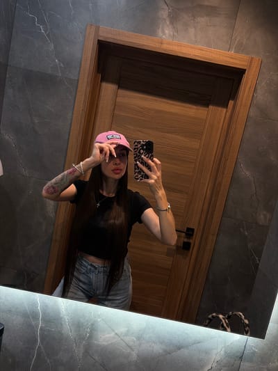 In a modern bathroom, a woman is taking a mirror selfie. She is wearing a pink baseball cap, a black t-shirt, and denim shorts. She has long dark hair and is wearing jewelry, including bracelets and a cross necklace. She is also holding a smartphone with a leopard print case. The bathroom features dark marble walls and a wooden door. The overall lighting suggests it is indoors, likely in a residential or hotel setting. There is no specific information about the location being in Dnp Ryzhiki, Russia.