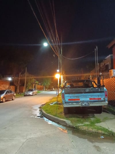 The image shows a nighttime scene in El Talar, Argentina, at the coordinates -34.472 latitude and -58.64 longitude. A blue pickup truck is parked on the right side of the street, facing away from the viewer. The truck has a license plate that reads "TG245." There are several power lines running across the street, illuminated by a streetlight on the left side of the road. The street is asphalt and there is a curb on the right side. There are trees on the left side of the road. There are two other cars parked on the street in the distance. There are no people visible. The street is relatively empty with minimal traffic. The scene is dimly lit and the sky is dark. The image is likely captured at night, possibly at a time when most people are not out on the streets.