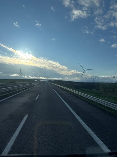 The image displays a multi-lane asphalt highway in Carapelle, Italy, under a blue sky with scattered white clouds and direct sunlight originating from the left. The road features white solid and broken lane markings extending into the distant horizon. Silver metal guardrails line both sides of the highway. Beyond the guardrails, green fields and sparse vegetation are observable. Multiple large wind turbines are positioned in the distant landscape on both sides of the highway, with a prominent turbine on the left. A concrete overpass spans the highway in the mid-ground. No individuals or active vehicles are present within the visible area.