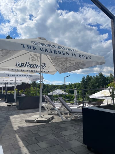 The image captures an outdoor seating area with a view of trees under a partly cloudy sky. In the foreground, several white lounge chairs are arranged on a paved patio. A large white umbrella with the text "THE GAARDEN IS OPEN" is prominent. Behind the lounge chairs, black planters are visible. In the background, a railing separates the seating area from a natural landscape with dense trees. The scene suggests a relaxed atmosphere, possibly at a cafe or resort in Zazymia, Ukraine.
