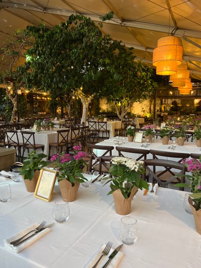 The image depicts a meticulously set outdoor dining area under a tent, likely for an event. The scene is located in Beit Hanan, Israel. White tablecloths cover several round tables, each adorned with place settings of silverware, napkins, and glassware. Small potted plants with pink and white flowers serve as centerpieces, some accompanied by framed table numbers, such as "1" and "2". Dark wooden chairs with woven seats are arranged around the tables. Lush green trees with dense foliage, illuminated by strings of warm fairy lights and overhead pendant lamps, create a natural and intimate ambiance. The lighting suggests it is evening or dusk. The overall impression is one of an elegantly prepared outdoor venue for a gathering or celebration.