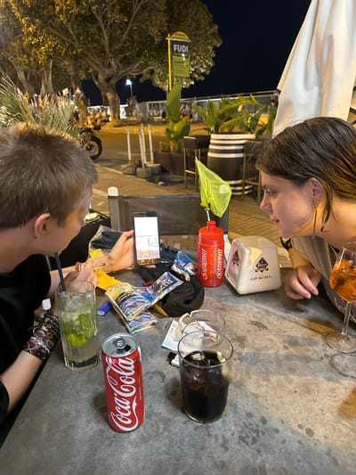 The image depicts an outdoor table setting at night, likely a cafe or restaurant, in Ospedaletti, Italy. Two individuals are seated at the table. On the left, a person with short blond hair is looking at a smartphone, holding a cigarette. In front of them is a glass with a green beverage containing ice and mint, a red Coca-Cola can, and packets of tobacco products. On the right, a person with dark hair is looking towards the smartphone. A white napkin dispenser with Italian writing ("Caffè dal 1863...") and a red water bottle are also on the table, along with an ashtray and a glass of dark liquid with ice. In the background, trees, a motorcycle, and other patrons are visible. The overall atmosphere suggests a casual evening gathering.