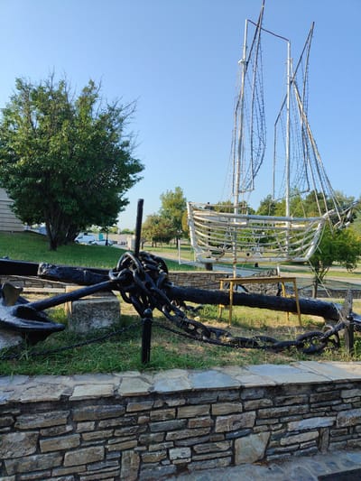 In Litochoro, Greece, a large, black, weathered anchor lies horizontally in the foreground, secured by a thick, rusty chain. The anchor is positioned on a bed of green grass, partially resting on a rough stone block. Behind the anchor, a metal fence with a black ball-topped post runs horizontally, separating the anchor from the rest of the scene. Beyond the fence, a large, abstract sculpture of a sailing ship, made of metal framing and adorned with what appear to be lights, stands tall against a clear blue sky. The ship sculpture's hull is outlined with white metal, and its masts are constructed of thin poles. To the left of the anchor and ship sculpture, a large, leafy green tree with visible branches stands prominently. Further in the background, more trees and greenery are visible, along with a light-colored building and parked vehicles, suggesting an urban or park-like setting. The ground is a mix of grass and paved areas.