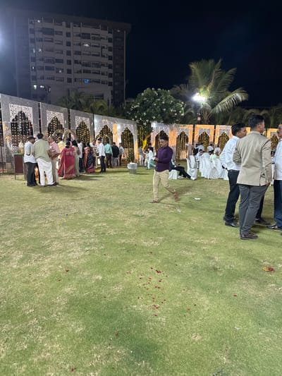 This photo depicts a group of people at an outdoor event at night in Nashik, India. They are standing on a grassy lawn next to a decorated structure with lights. Some are dressed in formal attire. A tall building is visible in the background.