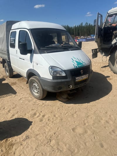 A white van with a gray tarp-covered cargo bed is parked on a sandy surface, likely in Чикшино, Russia. The van has a license plate reading "M459BT 164 RUS". A logo with turquoise geometric shapes and the text "hsk.ru" is visible on the front of the van. A dog is partially visible underneath the front of the van. In the background, a tall, dark-colored vehicle with its door open is parked next to the van. Beyond that, there are trees, tents, and possibly event structures set up on the sandy terrain under a clear blue sky with a few scattered clouds. The ground is covered in sand with visible tire tracks and footprints.