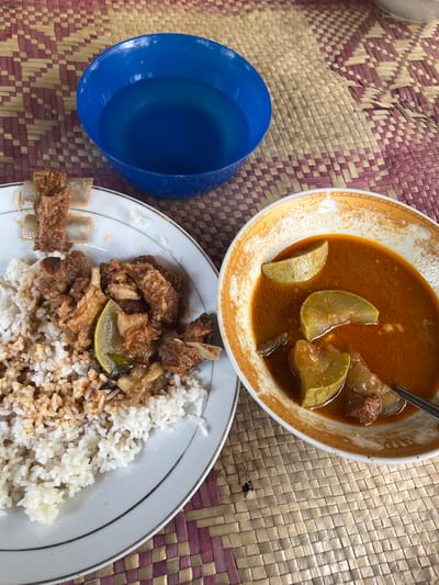 A close-up, overhead shot captures a meal set on a woven mat, likely in a home in Peurupok, Indonesia. The mat, with its alternating patterns of light tan and pinkish-purple, forms the backdrop. To the upper left, a bright blue bowl is filled with clear liquid. Dominating the center-left is a white plate piled high with fluffy white rice. Scattered over the rice are pieces of dark, savory meat, appearing to be ribs or braised chunks, with a slice of lemon nestled among them. To the right of the rice plate sits a decorative, orange-rimmed bowl overflowing with a rich, reddish-brown stew. Chunks of pale green vegetables, possibly chayote or zucchini, are visible submerged in the flavorful broth, along with more pieces of meat. A silver spoon rests in the stew, its handle partially submerged. The overall impression is of a simple, home-cooked meal, presented in a casual setting.