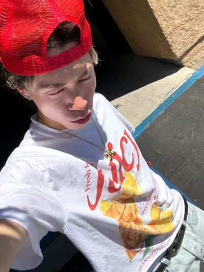 A person is lying down outdoors, possibly in a partially shaded area, wearing a red baseball cap, a white t-shirt with a red and yellow design, and a gold chain necklace. They appear to be relaxed, with their eyes closed. The background includes a concrete structure with a blue trim and a glimpse of what looks like denim clothing with a black belt. The location appears to be in the city of Indian Wells, United States of America.