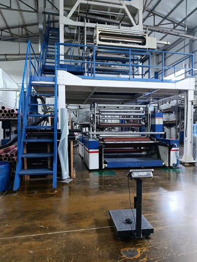 This is a photograph of the interior of a factory in the city of Oral, Kazakhstan, likely a plastic film manufacturing facility. The scene is dominated by a large, multi-level industrial machine, painted in blue and white with red accents. The machine features numerous rollers, belts, and structural components, suggesting it is used for producing or processing plastic film.
To the left of the main machine, a blue metal staircase with handrails leads up to a platform. Below the staircase, stacked cylindrical rolls of material, some with red and white banding, are visible. A blue barrel is also present on the left side.
In the foreground, a digital scale with a black platform and a vertical support pole is positioned on the factory floor. The floor itself is a dark, reflective surface with numerous scuffs and stains, indicating industrial use.
The background of the factory shows a high ceiling with exposed beams and lighting fixtures, typical of an industrial building. Shelving units holding white bags and more cylindrical rolls are visible in the distance, along with other pieces of machinery and equipment. A water cooler is also present on the right side. The overall impression is one of an active, but potentially older, manufacturing environment. There are no people visible in the image.