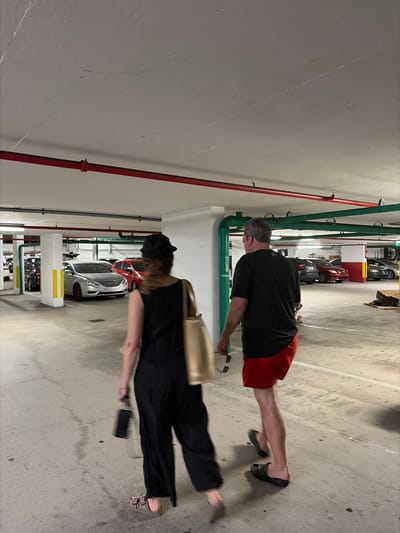 The image depicts two individuals walking in what appears to be a parking garage in Key Biscayne, United States of America. The first individual, closer to the camera, is wearing a black outfit and sandals, carrying a gold bag and a phone. The second individual, further away, is wearing a black shirt and red shorts, with black sandals. Various cars are parked in the background. The garage features overhead pipes, some painted red and green. The lighting is somewhat dim.