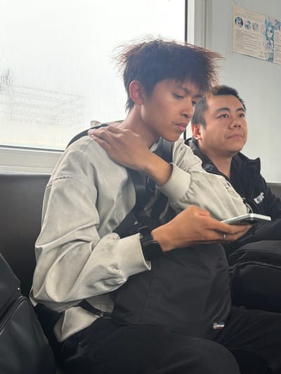 Two young men are seated closely together, likely in a public transport setting or waiting area, as suggested by the window and utilitarian seating. The image is oriented vertically, showing the subjects reclining horizontally.

In the foreground, one man with spiky dark brown hair and freckles is seen in profile, looking slightly upwards with a calm expression. He wears a light-colored long-sleeved top or jacket and has a black wristband or watch on his left wrist. His right arm is draped around the shoulder and neck of the second man.

The second man, positioned slightly behind and to the left of the first, has short dark hair and is looking upwards. He is wearing a black jacket over a light top. He holds a silver smartphone in his left hand, on which a ring is visible. His right arm is wrapped around the first man's waist or back, creating an embracing posture between the two.

The background includes a window showing a blurred, possibly wet or rainy exterior, indicating overcast weather. A piece of paper with text and diagrams is taped to the wall or ceiling above them, though the text is unreadable. A small rectangular tag on the second man's black apparel or bag clearly displays "NBA Classic." Dark bags or straps are visible, reinforcing a travel context. The overall scene suggests a moment of rest, intimacy, or comfort during travel. While the provided context is Nizhneleninskoe, Russia, the visual cues within the image are generic for travel.