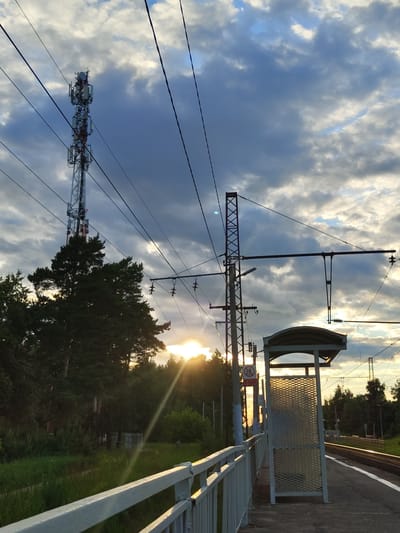 This image depicts a sunset scene at a train station in Tsibino, Russia. The sky is filled with dramatic clouds, with the sun setting behind a cluster of trees on the left, casting a warm glow and sunbeams across the scene. In the foreground, a white metal railing lines the edge of the train platform. To the right of the railing is a bus stop shelter with a curved roof and a mesh-like side. The platform itself is visible, with train tracks running alongside it. A tall cell tower stands prominently in the background to the left, towering over the trees. Electrical wires crisscross the sky above the platform and tracks. The overall environment appears to be a semi-rural or suburban area, with dense foliage surrounding the train station. There are no people visible in the image.