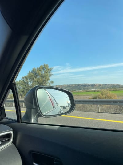 The image depicts an exterior view from the passenger side of a vehicle in motion on a highway near Jisr az-Zarqa, Israel. In the foreground, the dark interior of the car is visible, including a portion of the A-pillar on the upper left and the door panel with an air vent and handle on the lower left. The passenger side mirror, centrally positioned on the left, reflects part of the car's body and multiple vehicles traveling on the highway behind. Outside the car, a multi-lane asphalt highway is visible with a solid yellow line delineating a lane. A metal guardrail runs parallel to the road on the right. Beyond the guardrail, a green agricultural landscape with scattered trees, including a large tree with light green foliage on the left, extends towards the horizon. In the background, a long, light brown, rocky cliff formation or range of hills stretches across the scene under a clear blue sky marked by faint, wispy white clouds.
