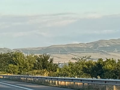This media file depicts a scenic landscape with a focus on a highway guardrail in the foreground and rolling hills in the background. The guardrail, made of metal with reflective elements, runs horizontally across the lower portion of the image, suggesting the perspective of someone traveling by road. Beyond the guardrail, a line of lush green trees forms a dense foliage barrier. The background is dominated by undulating hills, with visible terracing or paths cut into their slopes, possibly indicating agricultural activity or infrastructure. Several power lines or cables are also discernable stretching across the hills. The sky above is overcast with soft, diffused clouds, hinting at a temperate or possibly humid weather condition. The lighting suggests it is either early morning or late afternoon, as the light is gentle and shadow play is minimal. No people are visible in the image. The overall impression is one of a rural or semi-rural setting in Georgia, characterized by natural beauty and signs of human presence through the infrastructure. There is no visible text in the media file.
