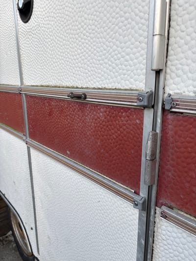 The image shows a close-up view of the exterior of a white and red caravan parked outdoors. The caravan has a textured white surface with horizontal red panels. Metal trim and hinges are visible along the seams and edges of the caravan's body and door. A black handle is partially visible at the top left of the frame. The ground beneath the caravan is not clearly visible, but appears to be an outdoor surface. The overall condition of the caravan suggests it is an older model. The image was taken in Kableshkovo, Bulgaria.