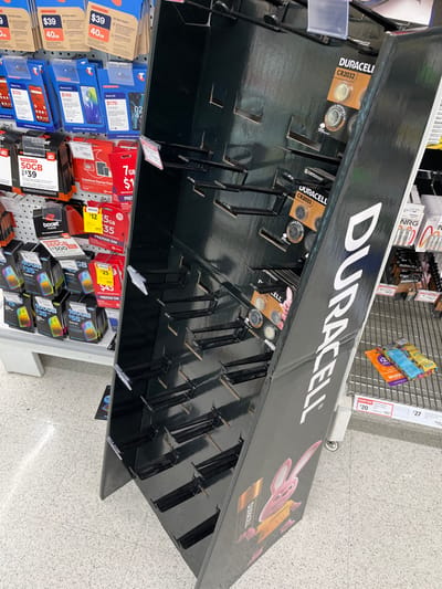 A Duracell battery display stand in a store, located at -27.555 latitude and 152.922 longitude, is mostly empty except for a few Duracell CR2032 batteries. The display is made of black cardboard and features a pink cartoon rabbit logo. The stand is located in a retail environment, possibly a supermarket or convenience store. The floor is tiled in white and grey. The image was likely taken from a slightly elevated angle, as the display is viewed from above. There are various items displayed around the Duracell display, including phone cards and accessories. The store is brightly lit, and the image is in focus. There are no people visible in the image.