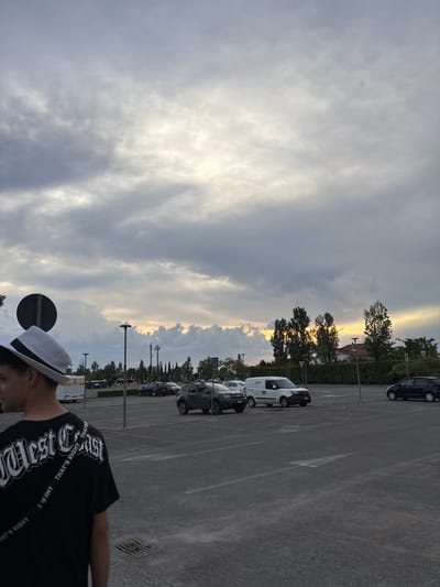 The image captures a parking lot scene in Pisa, Italy, under a cloudy sky with a sliver of golden sunset light breaking through. Cars are parked in rows, and trees line the perimeter. A person wearing a hat and a black t-shirt with white lettering is partially visible in the foreground. A street sign with a black circle on it is to the left of the person.