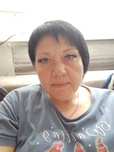 A woman with short black hair and blue eyes is taking a selfie indoors. She is wearing a light blue t-shirt with a gold chain necklace. Her expression is neutral. The background shows a textured wall and a sofa. The image appears to be taken during the daytime. There is no visible text in the image. The location is Pugachyov, Russia.