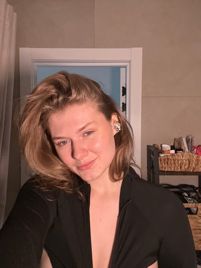 The image displays a female individual, appearing as a young adult, positioned indoors in what is identified as Yanga-Yul, Russia. She has fair skin, light-colored eyes, and medium-length light brown hair, which is styled to the side. The subject is looking directly at the camera with a subtle smile. She is dressed in a black, long-sleeved garment, possibly a top or jacket, with a front zipper partially open, exposing the upper chest area. A decorative ear cuff, appearing to be composed of small pearls and gold-toned accents, is visible on her right ear. The background shows a light-colored curtain on the left, a white doorframe enclosing a section of a light blue wall, and a beige, possibly tiled, wall segment on the right. To the far right, a dark shelving unit is present; its top shelf holds a wicker basket containing various small objects, including bottles and tubes, while darker items rest on a lower shelf. The scene is illuminated by interior artificial lighting.