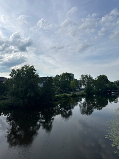 The image shows a body of water with a reflection of trees and buildings. The water is calm with some ripples. On the far bank, there are several trees and buildings. The sky is cloudy with some patches of blue. This scene is of the Lahn river in Wetzlar, Germany.