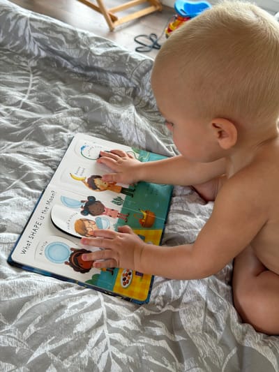 An infant with blonde hair is sitting on a bed with gray and white patterned sheets, holding a children's book. The book is open, displaying illustrations of characters and shapes, with text such as "What SHAPE is the Moon?" and "A circle." The infant's hands are touching the pages of the book. The environment appears to be indoors, possibly a bedroom. The location is not specified.