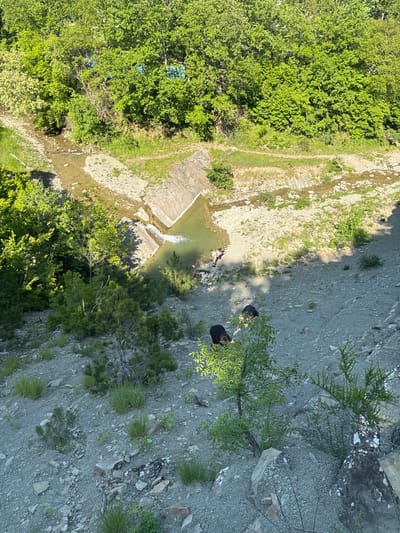 The image depicts a top-down view of a landscape near Kozichino, Bulgaria. A stream flows through a rocky riverbed, leading to a small dammed pool with a slight waterfall. Dense green vegetation covers one side of the stream. On the opposite side, a steep, rocky slope rises, featuring sparse vegetation. Two people are standing near the edge of the slope, close to a small tree. One individual is wearing black clothing, while the other has on a black shirt and lighter colored pants. The terrain consists of rock, gravel, and soil.