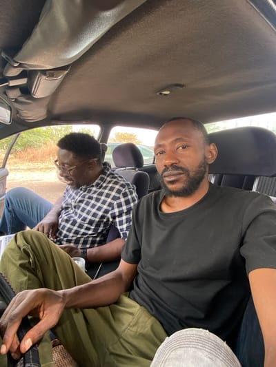 This image captures two adult males inside the front section of a vehicle, situated in Ungwan Liman, Nigeria.
The man in the foreground, positioned on the right, appears to be in the driver's seat. He has dark skin, short dark hair, and a full beard and mustache. He is wearing a plain black short-sleeved t-shirt and olive green cargo-style pants. His hands are resting in his lap, with the right hand partially obscured by a dark object, possibly the steering wheel. He looks directly at the camera with a neutral expression. A segment of a light-colored, possibly distressed denim-clad leg is visible in the lower right corner, belonging to an unseen individual or an unusual posture.
The second man is seated in the front passenger seat on the left. He has dark skin, short dark hair, and a trimmed beard or goatee. He wears dark-framed glasses, a dark blue and white checkered button-up shirt, and blue jeans. He is leaning forward, looking down at a white rectangular object, possibly documents or an electronic tablet, resting on his lap. A white cup is visible near his right hand, and a dark wristwatch is on his left wrist.
The vehicle's interior features a dark grey/brown fabric headliner. A sun visor and rearview mirror are visible on the left, along with an overhead console light fixture. The seat upholstery is dark and appears to have a subtle pattern. Through the windows, an outdoor environment with sparse, dry-looking vegetation and glimpses of other vehicles under daylight conditions can be seen.