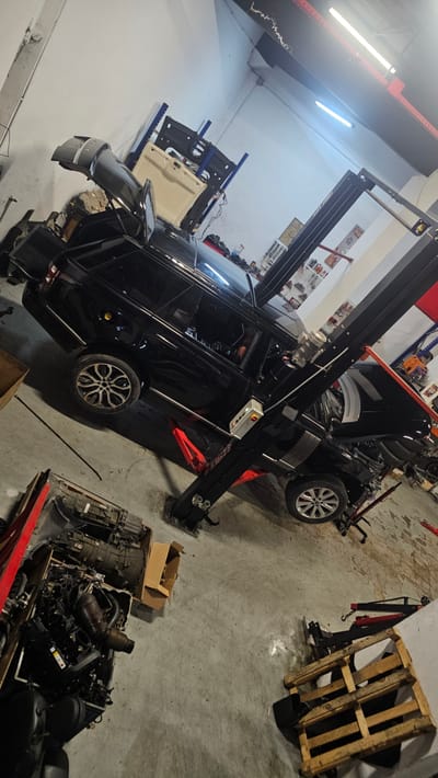 A black car is being worked on in a garage. The car is on a lift, and its hood is open. There are several parts of cars on the floor around it, including an engine. The garage is lit by fluorescent lights. There is a wooden pallet in the background. The scene suggests car repair or maintenance is taking place. The time of day is not discernible. The weather is not visible. Location cues such as "Ataşehir, Turkey" are provided, but not visible in the image. No people are visible in the image. No emotions are conveyed. No visible text is present in the image.