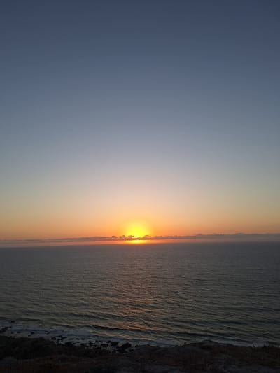 This image depicts a serene sunset scene on the coast of Ahrara, Morocco. The sky transitions from a deep blue on the left to a warm orange and yellow gradient as it nears the horizon, indicating it is either sunrise or sunset. The sun itself, a brilliant orb of yellow and orange, is partially obscured by the horizon line, casting a golden glow across the sky.

The scene is dominated by the vast expanse of the ocean, which occupies the right half of the frame. Gentle waves with white crests roll towards the shore, creating a textured surface on the water. Along the right edge, a dark, rocky coastline is visible, with some areas of dark sand or pebbles. The water appears calm, with the light reflecting off its surface, adding to the tranquil atmosphere.

There are no visible people or man-made objects in the frame, emphasizing the natural beauty of the location. The overall mood is peaceful and contemplative, inviting the viewer to appreciate the natural spectacle of a coastal sunset. The image suggests a clear, calm day, with no clouds present in the sky except for a faint line near the horizon. The time of day is definitively either dawn or dusk due to the intense colors in the sky and the sun's position near the horizon.