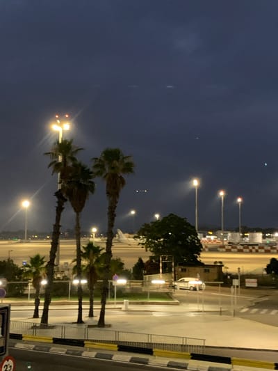The image depicts an outdoor scene at dusk, likely at an airport due to the presence of a runway, lights, and a distant airplane. The foreground features several palm trees, with their silhouettes stark against the darkening sky. A street lamp with bright lights stands prominently near the trees. In the middle ground, a white car is visible on a road, and beyond that, the airport tarmac with various buildings and checkered barriers. The sky is a deep blue-gray, with some faint lights and potential atmospheric phenomena visible. A traffic sign with the number "23" is partially visible in the lower left corner. This location is identified as Manises, Spain.