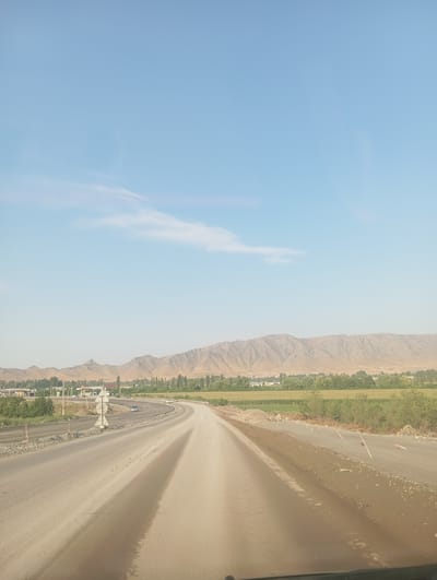 The image depicts a road under construction in the city of Жаны-Кыштак, Kyrgyzstan. The road is made of dirt and gravel, and appears to be a work in progress, with construction materials and barriers visible along the sides. In the background, there are mountains and trees, suggesting a rural or semi-rural setting. The sky is clear and blue. There are a few cars on the road, and some buildings are visible in the distance.