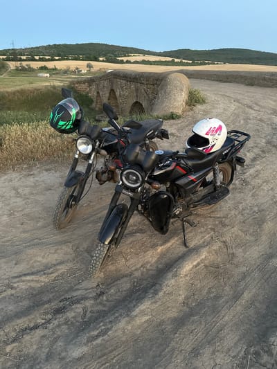 Two black motorcycles are parked side-by-side on a dirt road. The motorcycle in the foreground has a white helmet with pink and red markings on its seat. The motorcycle behind it has a green and black helmet hanging from its handlebars. In the background, there is an old stone bridge with arches, and a gently rolling landscape with dry grass and trees. The sky is clear and blue, indicating it is either early morning or late evening. The scene appears to be in a rural area, possibly the city of Filipeni, Moldova, given the context provided.