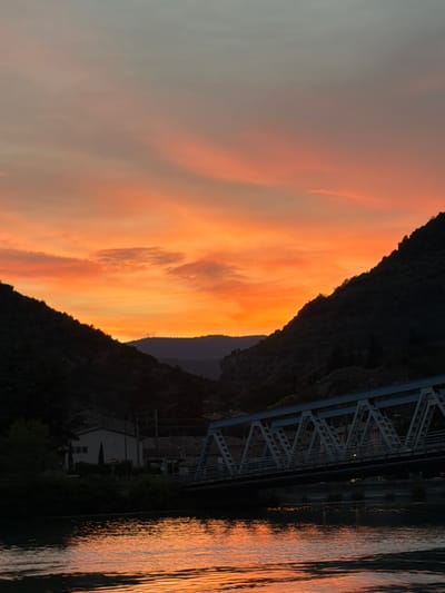 This image depicts a sunset over a river or body of water, likely in Loriol-sur-Drôme, France. The sky is a dramatic display of orange and pink clouds, transitioning to a soft gray at the top. A dark, silhouetted mountain range forms the backdrop on the left side of the image. A bridge, characterized by its metal truss structure, spans across the scene on the right. The river reflects the vibrant colors of the sunset, creating shimmering patterns on its surface. On the far right bank, some buildings and trees are visible, partially obscured by darkness. The overall mood is serene and picturesque, characteristic of a sunset. The time of day is clearly evening, and the weather appears clear, allowing for the vibrant sunset hues. No text is visible in the image.