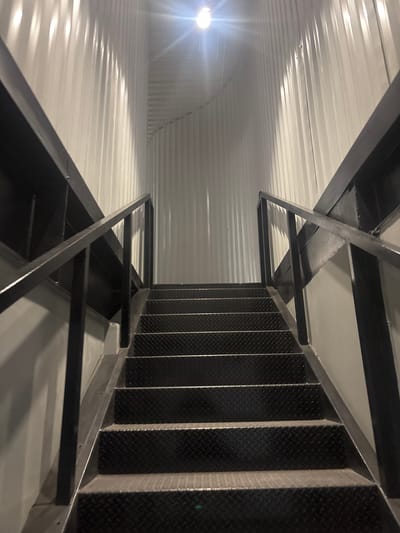 This image captures an upward view of a stark, industrial staircase within an indoor setting, likely in Buenavista, Mexico. The primary subjects are a dark metal staircase and a corrugated metal wall or ceiling.
The stairs are constructed from dark, possibly black, metal with textured diamond plate treads, indicating an industrial or utility design. They ascend steeply, supported by a sturdy framework of dark metal beams and angled braces that also form the surrounding structure. The handrails are integrated into this metal framework.
To the left, a corrugated metal surface, painted white or light grey, extends into the background. This surface is brightly illuminated by a very strong, unshielded light source positioned in the lower-left. The light creates prominent reflections and shadows that accentuate the ribbed pattern of the corrugated material, and a noticeable lens flare is present. The contrast between the bright, reflective wall/ceiling and the darker, textured stairs is a notable visual element.
No people or activities are visible, lending a sense of quiet functionality to the scene. The intense artificial lighting suggests an interior space, likely without natural light, such as a warehouse, factory, or commercial building. The overall aesthetic is utilitarian and robust. There is no discernible text in the image.