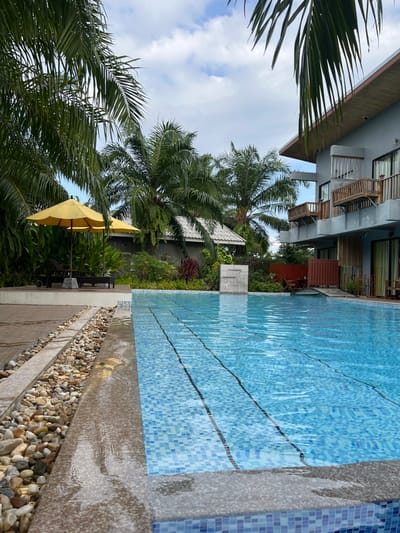 The scene depicts an outdoor swimming pool area in Ban Tha Muang, Thailand, under an overcast sky with patches of blue. The rectangular swimming pool, lined with light blue and white mosaic tiles, occupies the foreground and midground, containing clear, rippled blue water with visible dark horizontal lines on its floor. A dark grey stone coping borders the pool, showing wetness.
To the left of the pool, a section of light-colored river stones separates the coping from a brown wooden deck. On this deck, two black lounge chairs are positioned beneath two open, yellow market umbrellas. Beyond the deck, lush tropical vegetation, including numerous palm trees and various green plants, forms a dense backdrop. Partially obscured by the foliage, a single-story building with a white tiled roof and dark walls is visible in the midground-left.
At the far end of the pool, a light-colored rectangular vertical concrete structure stands. To the right of the pool, a multi-story building with light grey walls and a sloped dark roof features multiple wooden balconies with railings. Windows and glass doors are present on its facade, with wooden slat accents on the lower levels. No individuals or active interactions are observed within the scene.