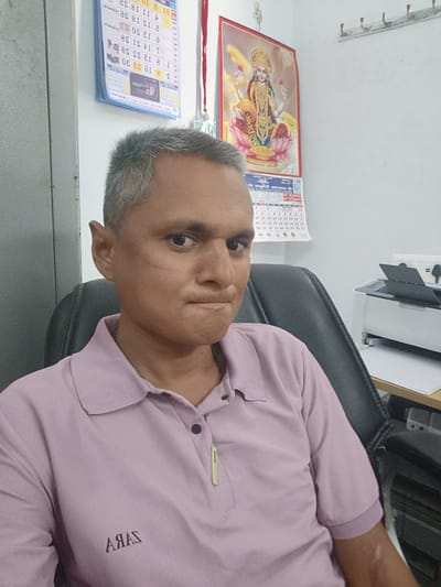 The image depicts a man sitting in a black office chair, wearing a pink polo shirt with "ARAS" printed on it. He has short, gray hair, and a scar is visible on his face. Behind him, there is a wall with two calendars and a poster of a Hindu goddess. A printer sits on a desk to the right. Based on the OCR data in the image, it might be from Dhandhuka, India.