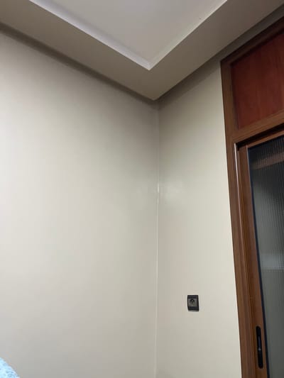 An interior view shows a window with a brown frame atop a light beige wall. A dark electrical outlet is near the top. The bottom of a white structure appears to the left. The wall has a smooth, reflective finish, suggesting recent painting. The composition is from a low angle, looking upwards.