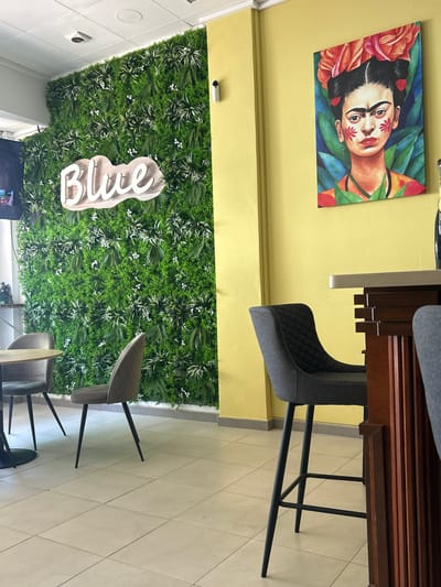 The image depicts the interior of a brightly lit establishment, possibly a cafe or restaurant, in Vera, Spain. A prominent feature is a large, textured green wall covered in artificial foliage and small white flowers, with a 3D wooden sign spelling "Blue" mounted on it. To the right of the green wall, a yellow-painted wall displays a vibrant painting of Frida Kahlo adorned with flowers. In the foreground, a dark grey, modern bar stool with a diamond-stitched pattern and thin black legs is positioned in front of a wooden counter. Two other similar stools are partially visible. The flooring consists of large, light-colored square tiles. Other visible objects include a camera mounted on the green wall, a table with chairs on the left side of the image, and part of a television screen on the far left. The scene is clean and well-maintained, suggesting a commercial space designed for patrons.