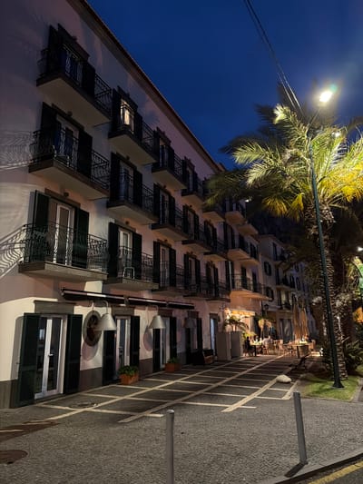The image shows a building at night in Ponta do Sol, Portugal. The building is light-colored, multistory, and has many balconies with black railings. Some balconies have planters. Streetlights illuminate the building and a palm tree adjacent to it, casting shadows on the facade. A street runs alongside the building, marked with white grid lines. In the background, there are tables and chairs, suggesting a restaurant or outdoor dining area. Some people are present in this dining area, but they are not clearly visible. The sky is a dark blue. The scene appears to be a quiet evening in the city.