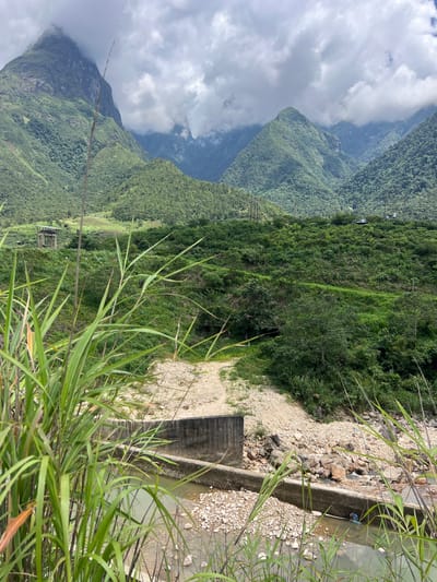 An overcast day frames a lush green mountainous landscape. In the foreground, tall green grass sways, obscuring a concrete structure and a shallow, rocky river. Beyond the river, the terrain rises into verdant hills covered in dense vegetation. A dirt path winds its way up the hillside. In the middle ground, a concrete observation tower stands on a grassy incline. In the distance, rugged mountains pierce the cloudy sky, their peaks partially shrouded in mist. A small, dark-colored structure, possibly a house or shed, is visible on a distant hillside. The overall scene depicts a natural, mountainous environment with elements of human infrastructure.