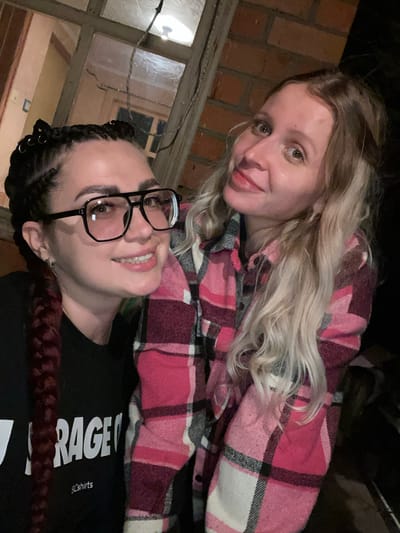 Two women are captured in a close-up, selfie-style photograph. The woman on the left has dark hair styled in braids adorned with small silver rings, wears large, dark-rimmed glasses, and a black t-shirt with white lettering. The woman on the right has long, blonde, wavy hair and is wearing a pink and black plaid shirt. Both women are smiling and appear to be outdoors, with a brick wall and a wooden window frame visible in the background. The lighting suggests it is dusk or nighttime. The location is identified as Солнечная Поляна, Russia.