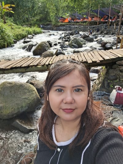 A person poses in a selfie next to a rocky stream. To the left, a wooden plank bridge crosses the water. Camping tents are visible in the background, suggesting a recreational area. The person is wearing a dark-colored top with white trim. The natural environment with the flowing water and vegetation adds to the scenic backdrop.