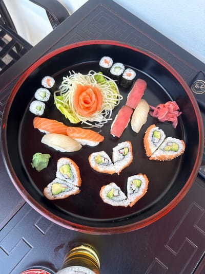 A visually appealing platter of assorted sushi and sashimi is the central focus of this image, presented on a large, round, black serving tray with a distinctive red rim. The tray rests on a dark, possibly wooden, table adorned with intricate, carved or embossed geometric patterns that suggest an Asian-inspired design.
The platter showcases a variety of Japanese delicacies. There are numerous pieces of nigiri, including vibrant orange salmon, deep red tuna, and pale white fish, each served on a bed of rice. Several types of maki rolls are present: smaller rolls containing salmon and cucumber, and larger inside-out rolls (uramaki) filled with avocado and crab stick, some featuring a topping of salmon. Many of these larger rolls are cut in half, revealing their fresh ingredients.
Traditional Japanese condiments are also included: a mound of pink pickled ginger (gari) is placed on the top left of the tray, and a dollop of green wasabi paste sits at the bottom right. A decorative element features thinly sliced salmon sashimi artfully arranged into a rose shape, presented on a bed of shredded white cabbage and a green lettuce leaf.
Beyond the platter, the top left corner of the image shows a black object, possibly a glasses case, with a discernible "Ray-Ban" logo. In the bottom right, a clear glass partially filled with a golden, foamy liquid (likely beer) is visible, next to a red bottle cap with faint, unreadable text. The scene is brightly illuminated by natural light, indicating a daytime setting. This meal is likely being enjoyed in a restaurant or home in Hainburg an der Donau, Austria.