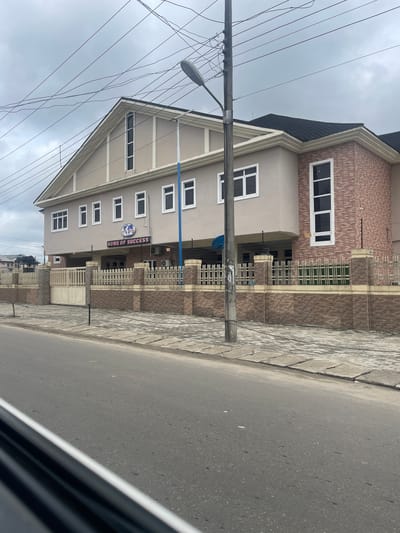 A two-story building with a dark pitched roof and beige exterior walls stands prominently on a street in Rumuokwuta, Nigeria. The building features numerous rectangular windows with white frames and a decorative brick facade on the right side. A prominent sign above the entrance reads "HOME OF SUCCESS" with a globe logo. The building is surrounded by a beige and brick fence with metal bars. A paved sidewalk and a road with faint markings are visible in the foreground. Overhead, a network of power lines and a street lamp are present. The sky is overcast with grey clouds. No people are visible in the image.