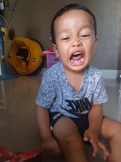 The image shows a young child with their mouth wide open, exhibiting a grimace. The child is wearing a grey t-shirt with a black Nike logo. They are sitting on a tiled floor. In the background, there is a yellow toy, a pink toy, and a blue fan. The scene appears to be indoors, possibly a home environment in Cibungbulang, Indonesia.