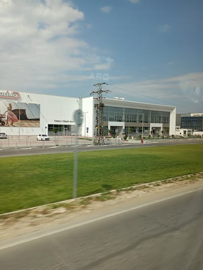 The image depicts a modern commercial building with a large white facade and extensive glass windows, situated in Tsaratsovo, Bulgaria. The building displays signage for "MOLINO" and "CERAMIC CENTER". In front of the building, there is a paved area with several parked cars, including a white hatchback and a silver sedan. A vibrant green lawn stretches across the foreground, bordered by a grey road and a light-colored, sandy strip. Overhead, power lines crisscross the sky, and a utility pole with electrical equipment stands prominently near the building. The sky is partly cloudy with patches of blue. The overall scene is a bright, daytime exterior shot of a commercial district.