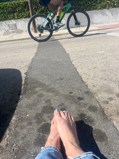 The image shows a section of paved road in La Marina De Creixell, Spain, with a bicycle on the left and a pair of bare feet on the right. The bicycle is turquoise with black wheels and appears to be resting on the side of the road, partially in shadow. The feet, with light blue nail polish, are also partially in shadow and appear to be resting on the road surface. A person wearing blue jeans is visible near the feet. The road surface is a mix of light and dark grey asphalt, with a visible seam or repair line running diagonally across the frame. There is greenery visible on the left edge of the image, suggesting the presence of vegetation along the roadside. The scene appears to be outdoors in daylight.