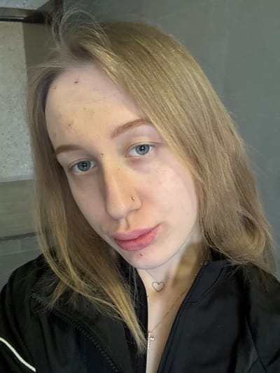 The image displays a close-up, slightly angled portrait of a young woman. She has fair skin, light blue eyes, and long, straight to wavy blonde hair that falls around her face and shoulders. Her eyebrows are light brown and well-defined. A small, metallic stud piercing is visible on her left nostril. Her lips are full and have a natural pinkish hue, showing some natural texture. Several small, reddish blemishes or marks are visible on her forehead, cheeks, and chin.
She is wearing a black zippered jacket or hoodie, visible across her chest and shoulder. A delicate, light-colored necklace featuring a small, sparkly cross-shaped pendant is worn around her neck. Below the necklace, on her upper chest, there is a small, outlined heart tattoo.
The background is an indistinct, plain light grey surface, consistent with an indoor setting, possibly a wall. The lighting is even and soft, suggesting an interior environment. The woman looks directly towards the viewer with a neutral to slightly contemplative expression. There is no visible text in the image. The location context for this image is Novoaleksandrovka, Russia.